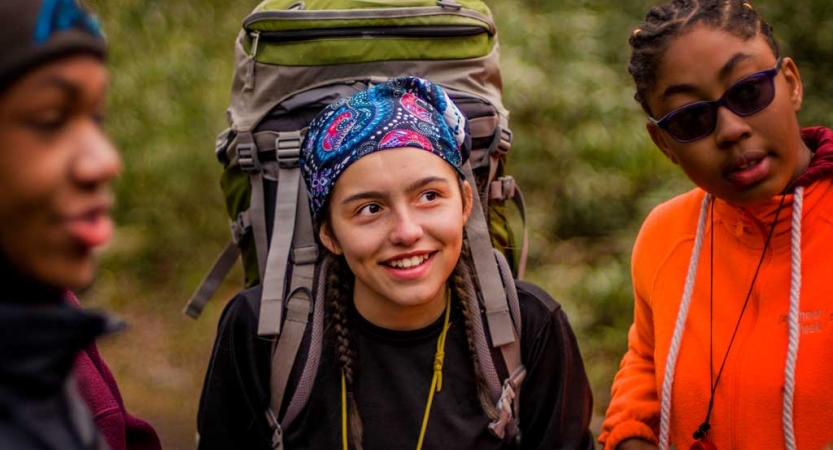 Three people stand in a green area. One of them wears a backpack. 